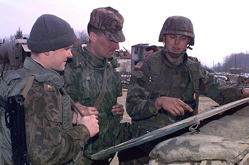 Three soldiers — two in Russian VSR three-colour woodland and one in U.S. woodland — lean over a map on a sandbagged position, 1996 Bosnia.