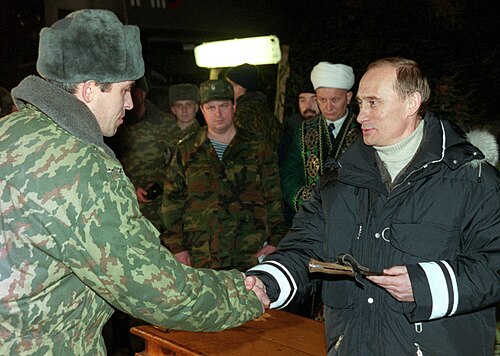 A civilian leader in a dark coat hands a decoration to a soldier while Russian soldiers in woodland/mottled camouflage stand in a dimly lit hall in Chechnya, 2000.