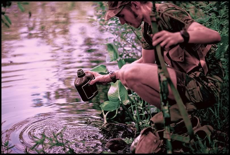 Modern re-enactor in Rhodesian Light Infantry 1977 kit, wearing Rhodesian Brushstroke camouflage, crouched by a river filling a water bottle