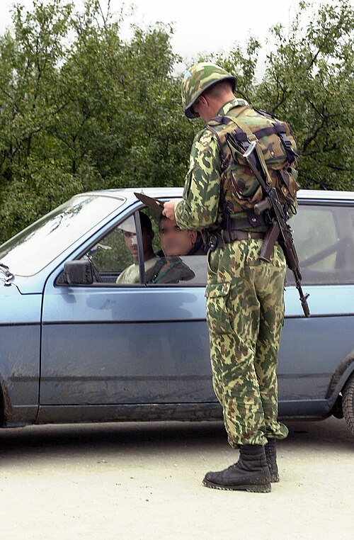 A Russian soldier in VSR/Dubok three-colour woodland camouflage and combat helmet, holding an AK-pattern rifle, speaking with a driver at a checkpoint