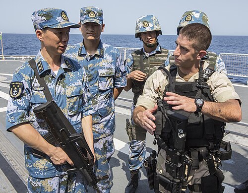 U.S. and Chinese Navy sailors talking on a ship deck, the Chinese crew wearing blue-green pixel Type 07 Ocean camouflage.