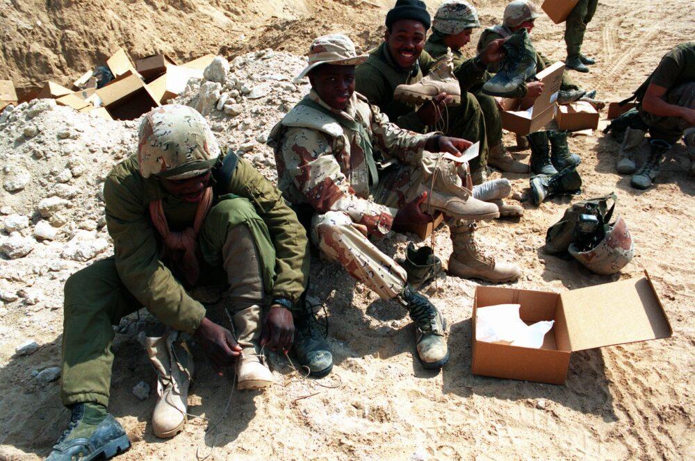 US Marines of C Company, 3rd Tank Battalion, 1st Marine Division trying on newly issued field boots in Saudi Arabia, 19 February 1991: one Marine wears the six-colour desert pattern on jacket and trousers, while another in the foreground is still in plain olive green.