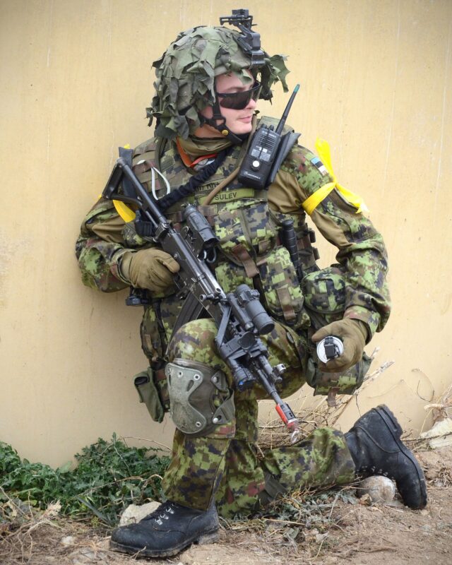 Estonian soldier in a pixelated woodland-pattern field uniform with a scrimmed helmet and a rifle, kneeling on a Spanish training area.