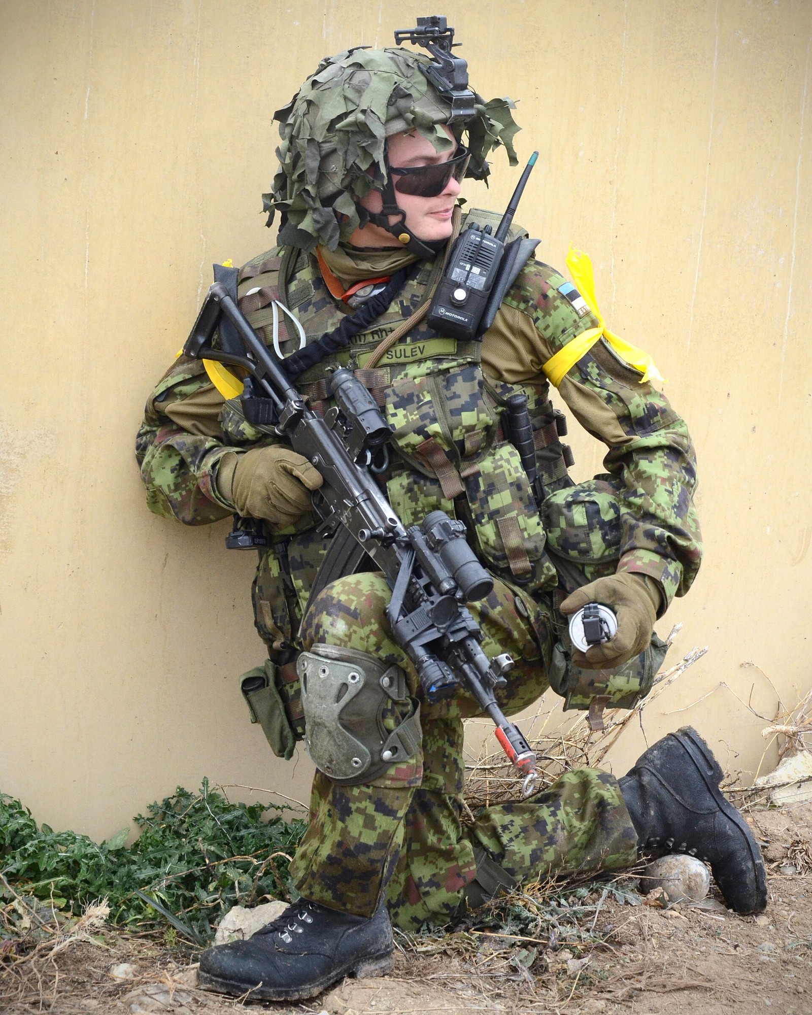 Estonian soldier in a pixelated woodland-pattern field uniform with a scrimmed helmet and a rifle, kneeling on a Spanish training area.