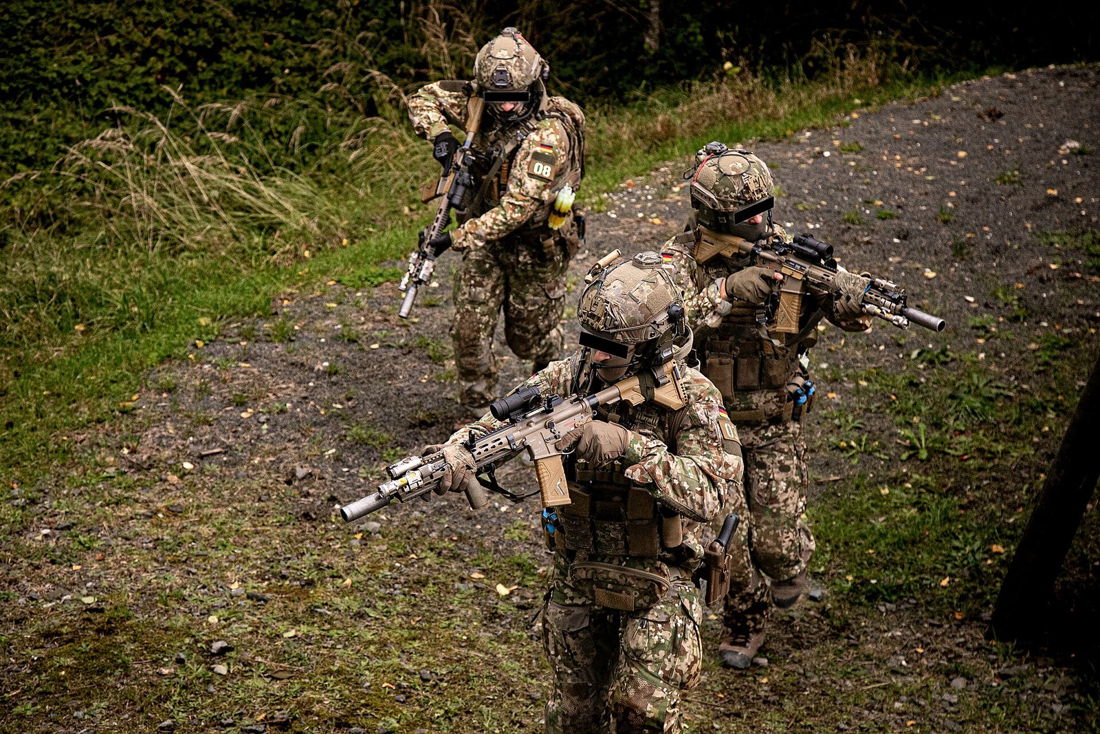 A column of German navy special forces operators in pale six-colour Multitarn camouflage walking in single file on a wooded track, wearing helmets and carrying suppressed rifles.