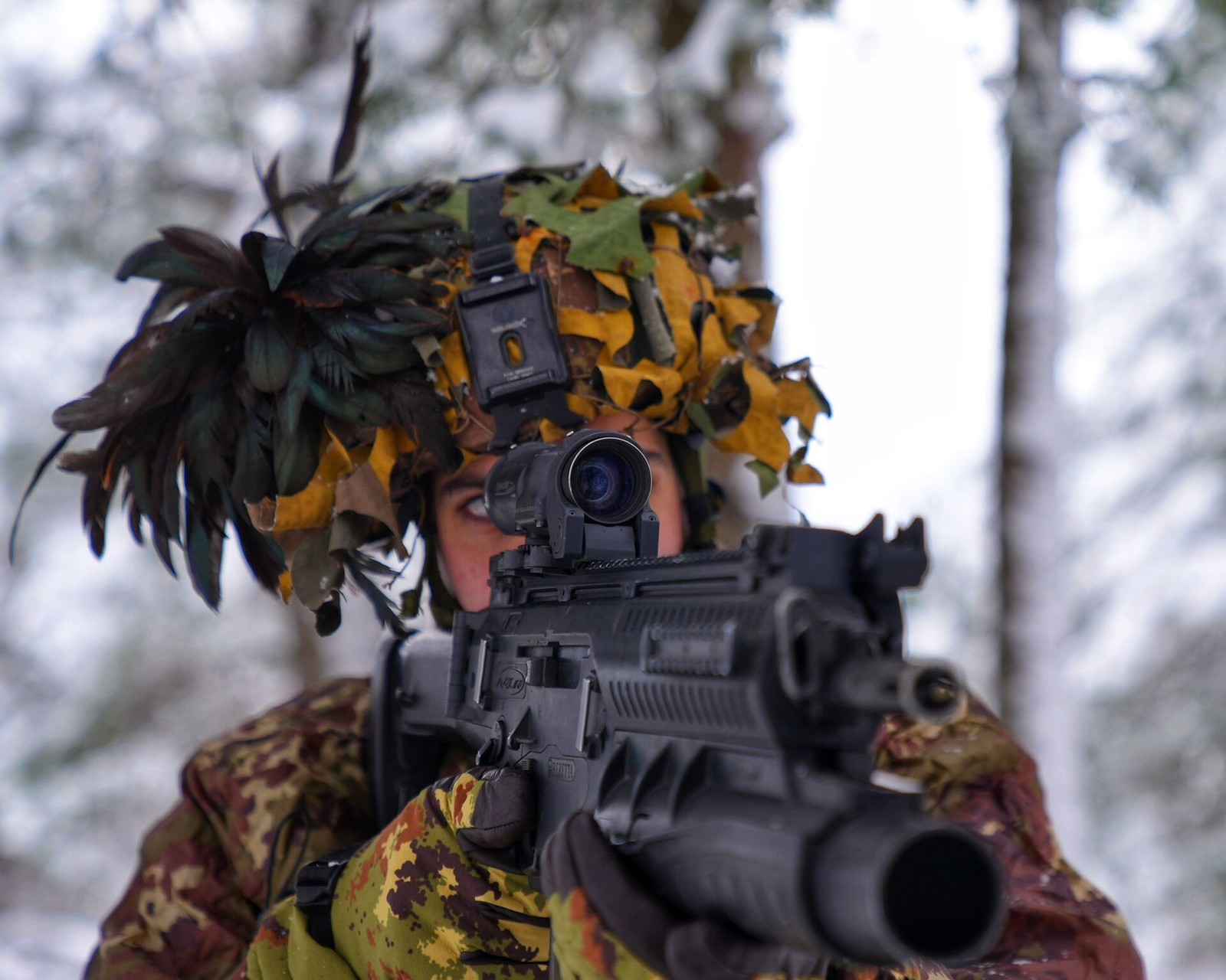 A single Italian soldier in green, brown and ochre camouflage uniform and matching helmet cover, seen from the chest up behind a modern assault rifle fitted with a grenade launcher and a sight, firing downrange in a snowy wood.