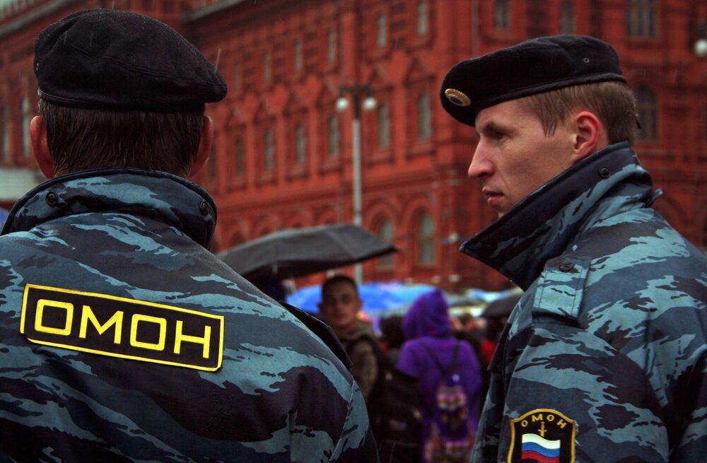 Two Russian police officers in dark blue wavy-striped camouflage, one seen from behind wearing a yellow Cyrillic OMON back panel, the other in profile, on a rainy day in front of a red brick building.