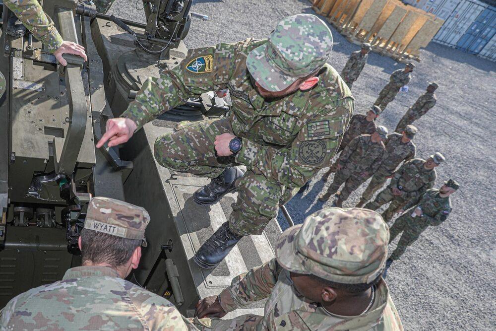 Slovak soldier in the blocky digital camouflage of the vz 2007 Les print crouched on the deck of a self-propelled artillery system and pointing down at its mechanism, watched by a group of U.S. soldiers in OCP uniforms.