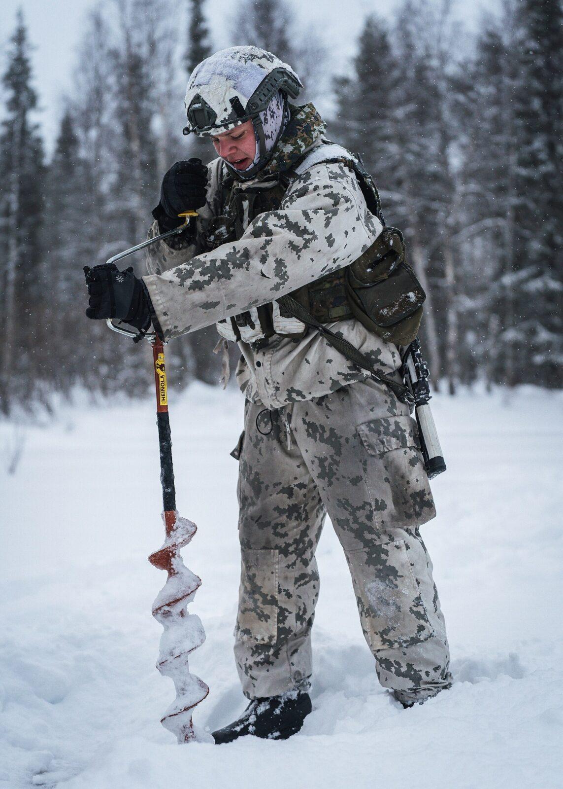 Soldier in an off-white snow camouflage suit with grey-green blotches, standing bent in deep snow and turning an ice auger with an orange handle, conifer forest and falling snow behind him.