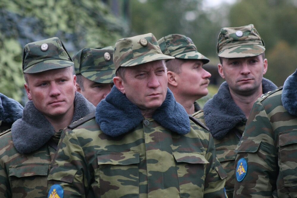 Men in banded green and tan camouflage jackets with fur collars and matching camouflage caps, standing outdoors in a line