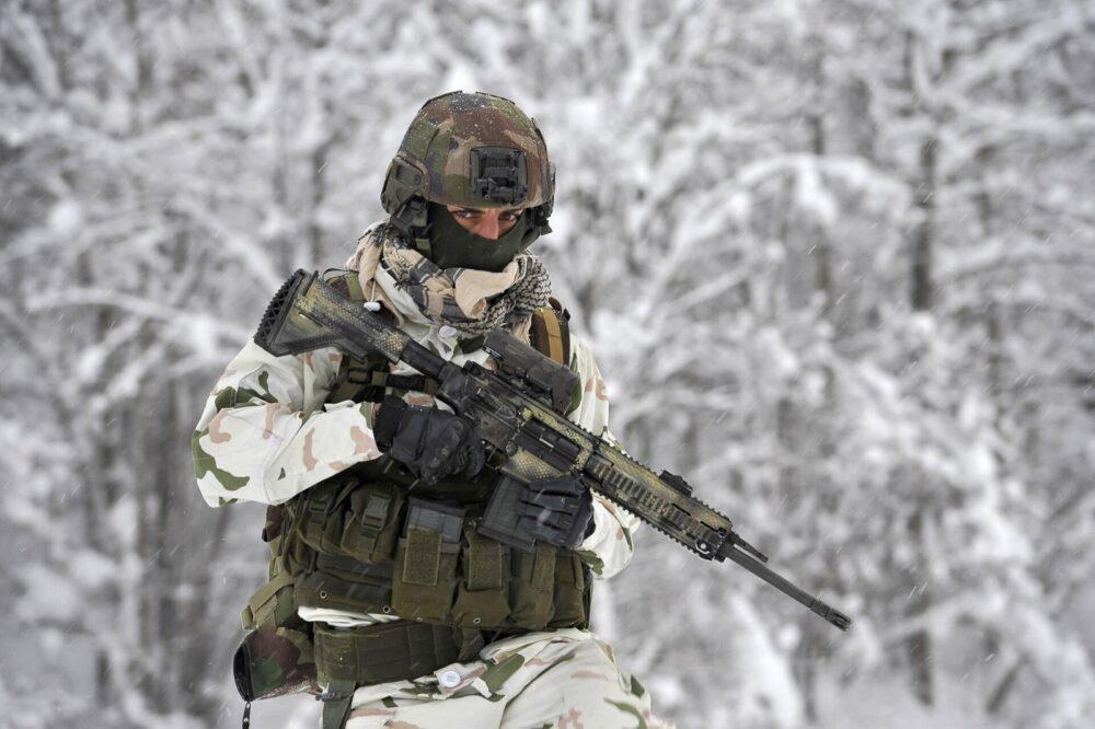 A soldier in pale snow camouflage — white ground with green and salmon shapes — wearing a dark green plate carrier and helmet, carrying a scoped rifle through fresh snow in a wood.