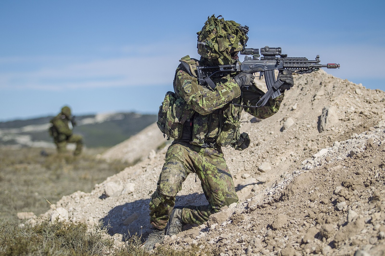 Estonian soldier kneeling in a pixelated green woodland camouflage uniform while aiming a rifle on a dry training area.