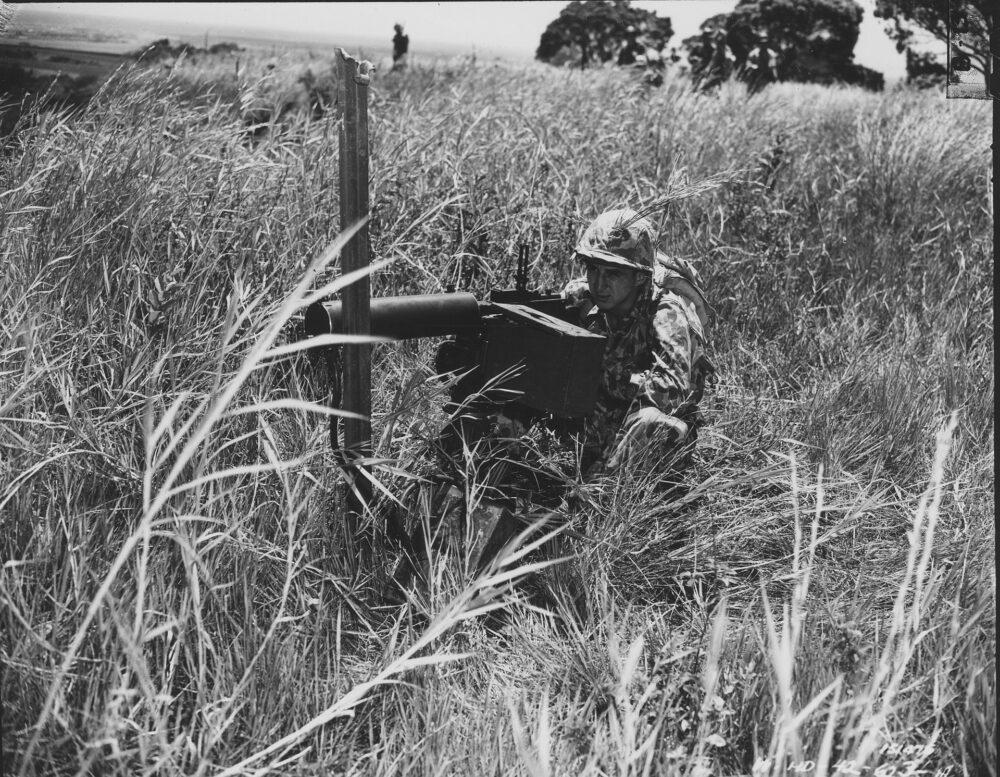A US Army soldier wearing 1942 camouflage uniform crouching in dense tropical vegetation for a concealment demonstration.