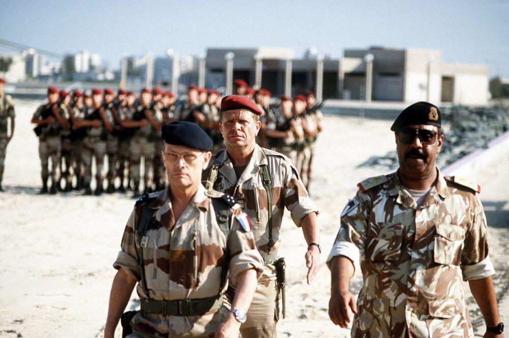 Three officers in desert uniforms walking abreast in front of a formed French detachment in Kuwait City, 30 March 1991: two in the wide-band French desert print and one, at right, in a lighter desert print with finer shapes.