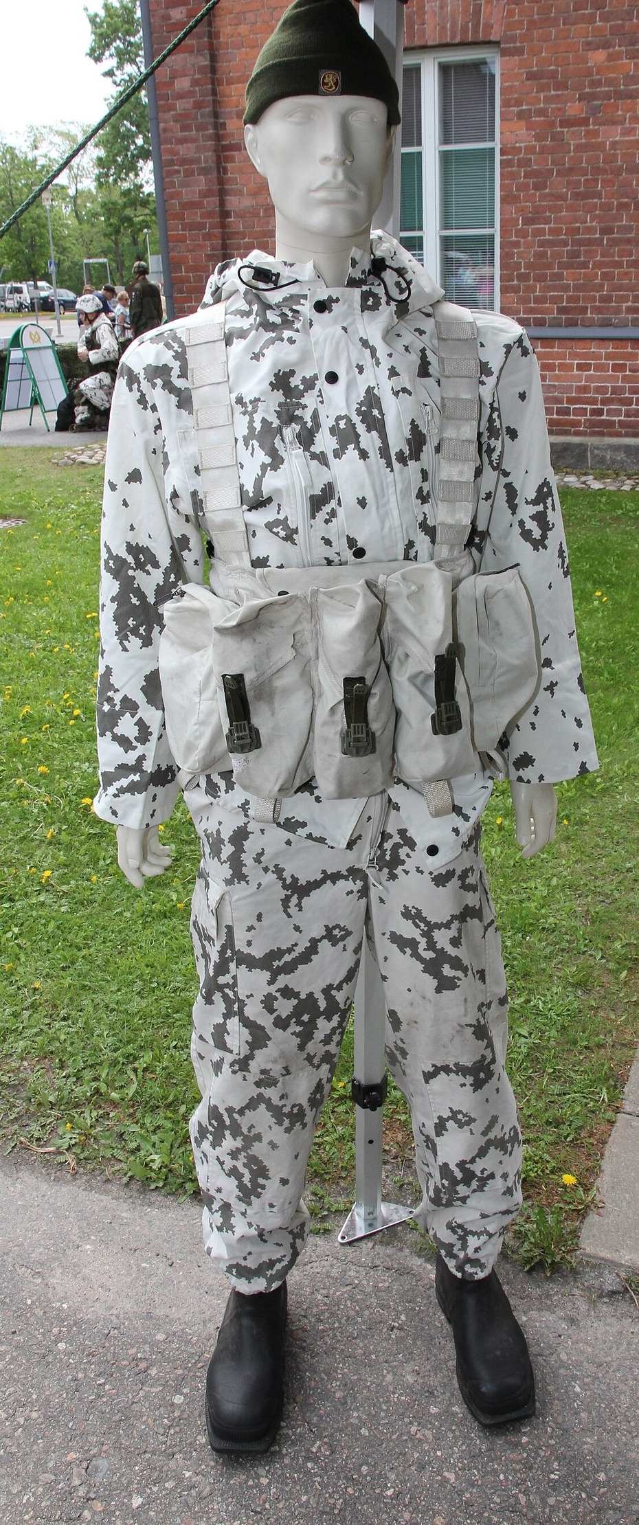 Display mannequin in a white snow camouflage jacket and trousers with dark grey-green blotches, wearing a green wool cap and light grey webbing with magazine pouches, outdoors in front of a red brick building.