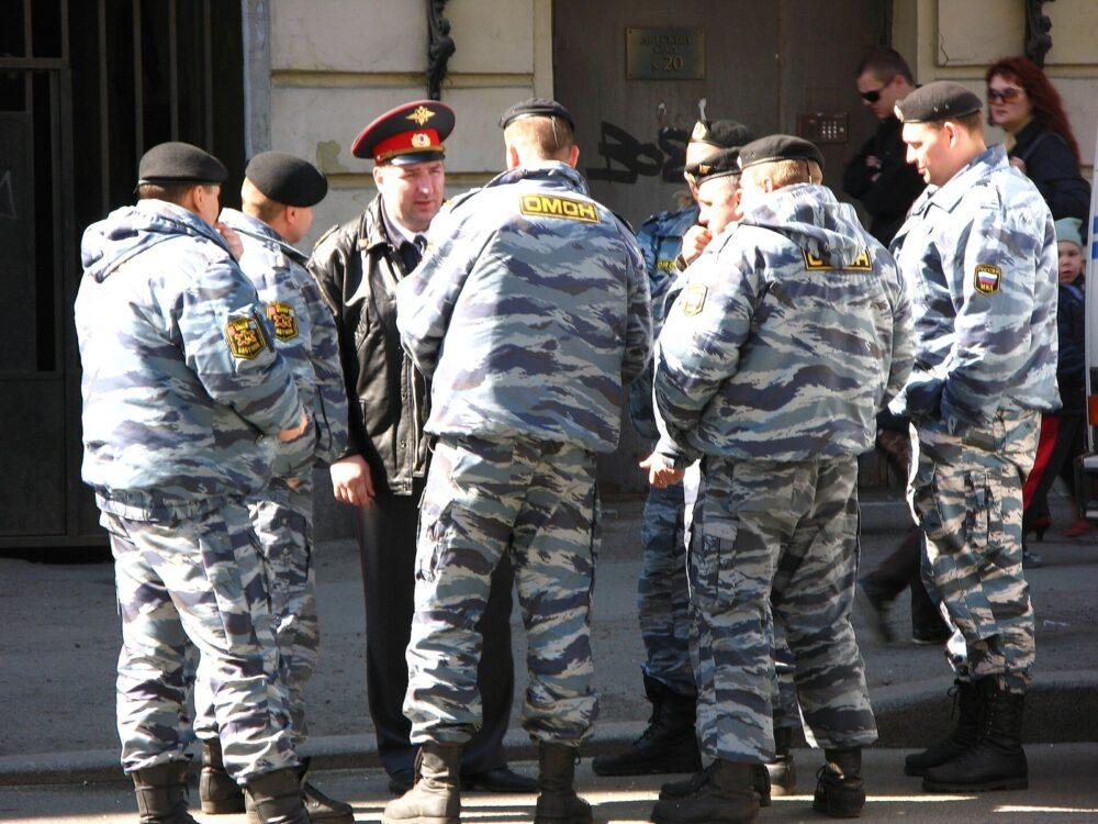A group of police officers in pale blue-grey wavy-striped camouflage jackets and trousers standing in a street around an officer in a black leather jacket and peaked cap, several with Cyrillic OMON panels on their backs.