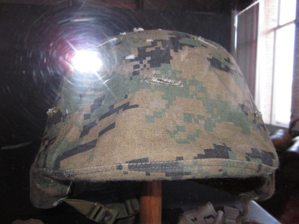 A military helmet with a blocky green and brown pixel camouflage cover, displayed on a stand in a museum case.