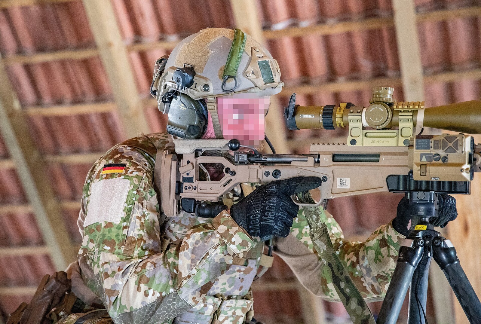 A German special forces sniper in Multitarn camouflage aiming a scoped rifle from behind cover inside a wooden building.