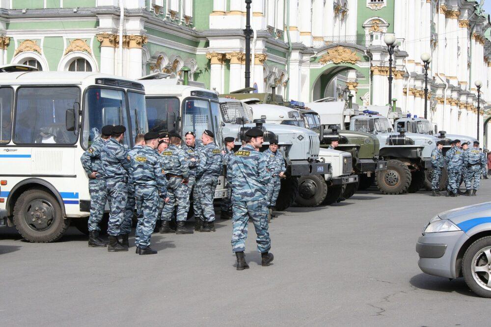 Police officers in pale blue-grey wavy-striped camouflage standing beside a white and blue police bus, with further white buses carrying a blue band and grey service vehicles parked behind them.