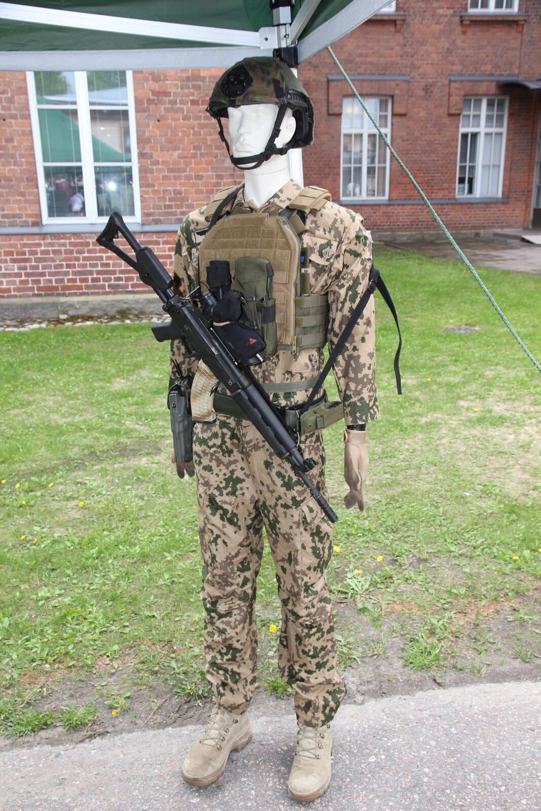 Standing display mannequin in sandy desert camouflage with green and brown blotches, body armour and webbing over the jacket, a helmet with a green camouflage cover, with a black assault rifle with a tubular optical sight slung across the body, a pistol in a holster at the hip.