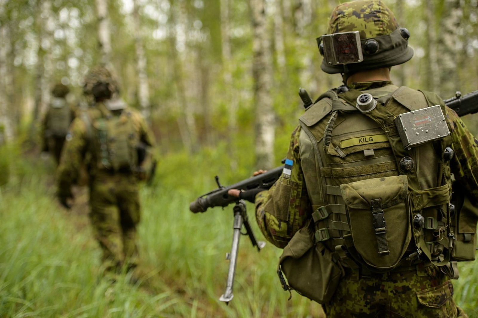 Estonian soldiers in pixelated camouflage walking through mixed woodland during a training lane in Latvia.