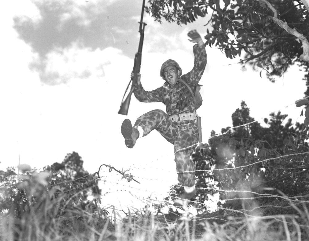 A US Marine Raider in camouflage uniform vaulting over barbed wire on a training obstacle course, his rifle held above his head.