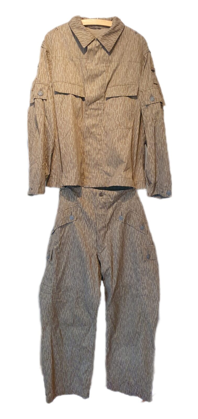 Field jacket and trousers of an East German Kampfanzug 64 hanging from a wooden hanger against a plain white background, printed in fine vertical brown dashes on a light khaki ground