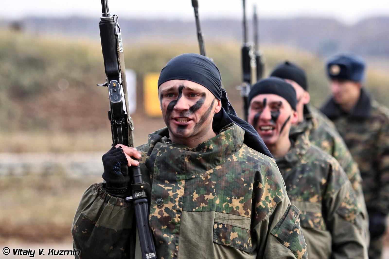 A Russian internal troops special purpose unit serviceman in camouflage uniform with black face paint holding an assault rifle, other servicemen out of focus behind him, at the Interpolitex 2010 exhibition, Moscow.