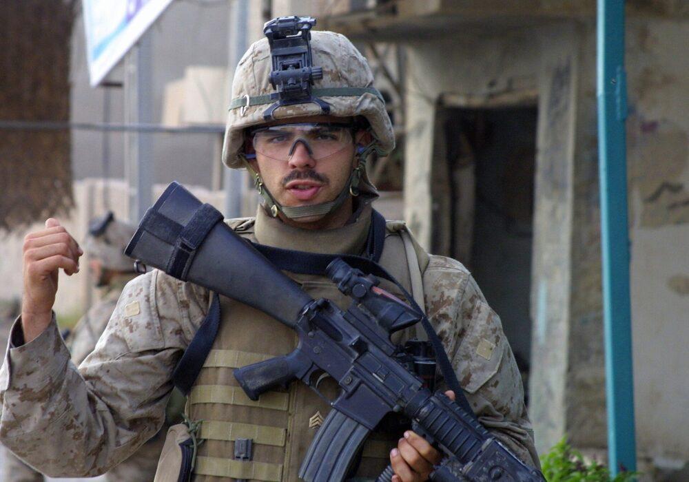 A U.S. Marine in a sandy pixel-patterned desert camouflage uniform and body armour, holding a rifle in a street in Iraq.