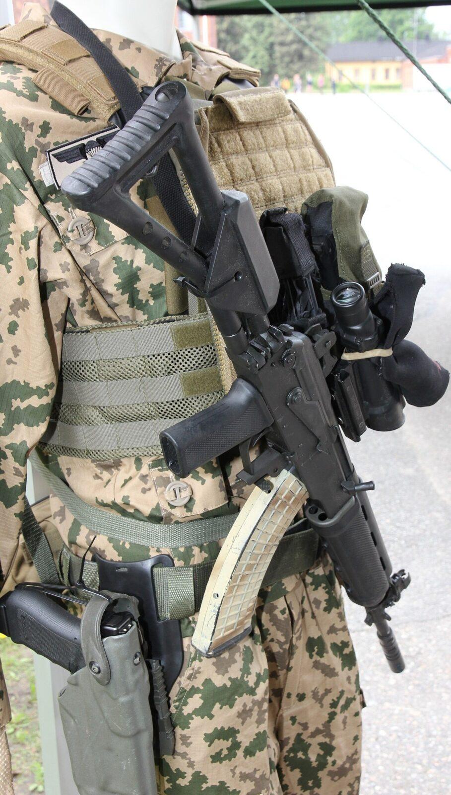 Close view of a desert-camouflage jacket and trousers, sandy ground with large green and brown blotches, under a tan body-armour vest with olive webbing, with a black assault rifle and a pistol holster crossing the frame.