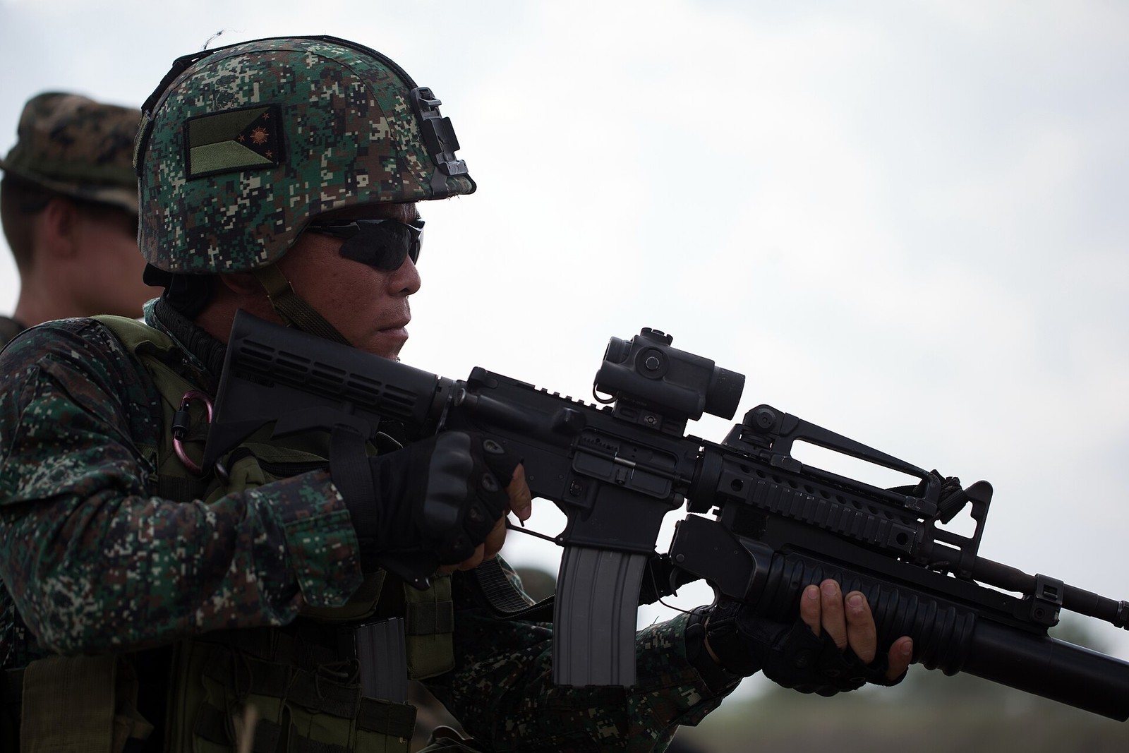 A Philippine Marine firing a rifle at a live-fire exercise, the green square-block digital camouflage clearly visible on his blouse and helmet cover, with an out-of-focus second figure in a green-and-brown patterned boonie hat at the left edge.