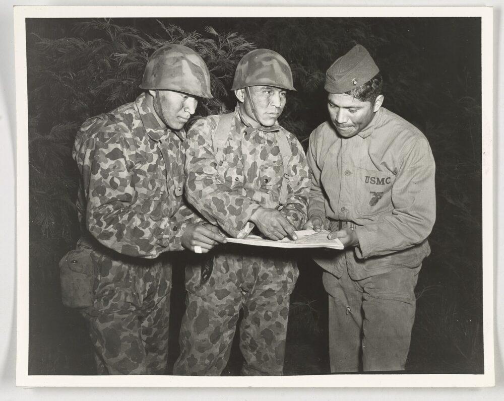 Two soldiers in camouflage uniform and one in plain uniform standing together over a document during a training exercise in the Pacific.