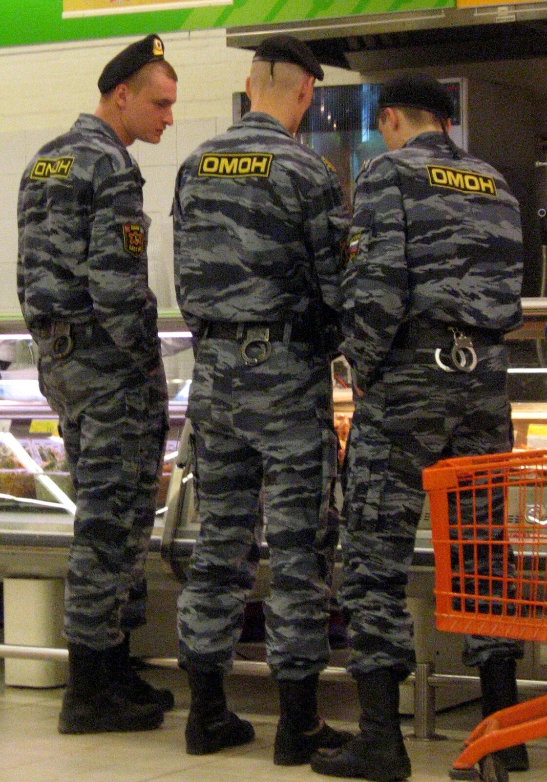 Three police officers photographed from behind at the glass display counters of a shop, wearing dark grey-blue wavy-striped camouflage jackets and trousers.