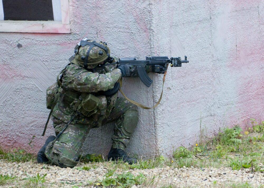 Kneeling soldier in blocky green-brown camouflage uniform and helmet, holding an assault rifle, in front of a concrete building.