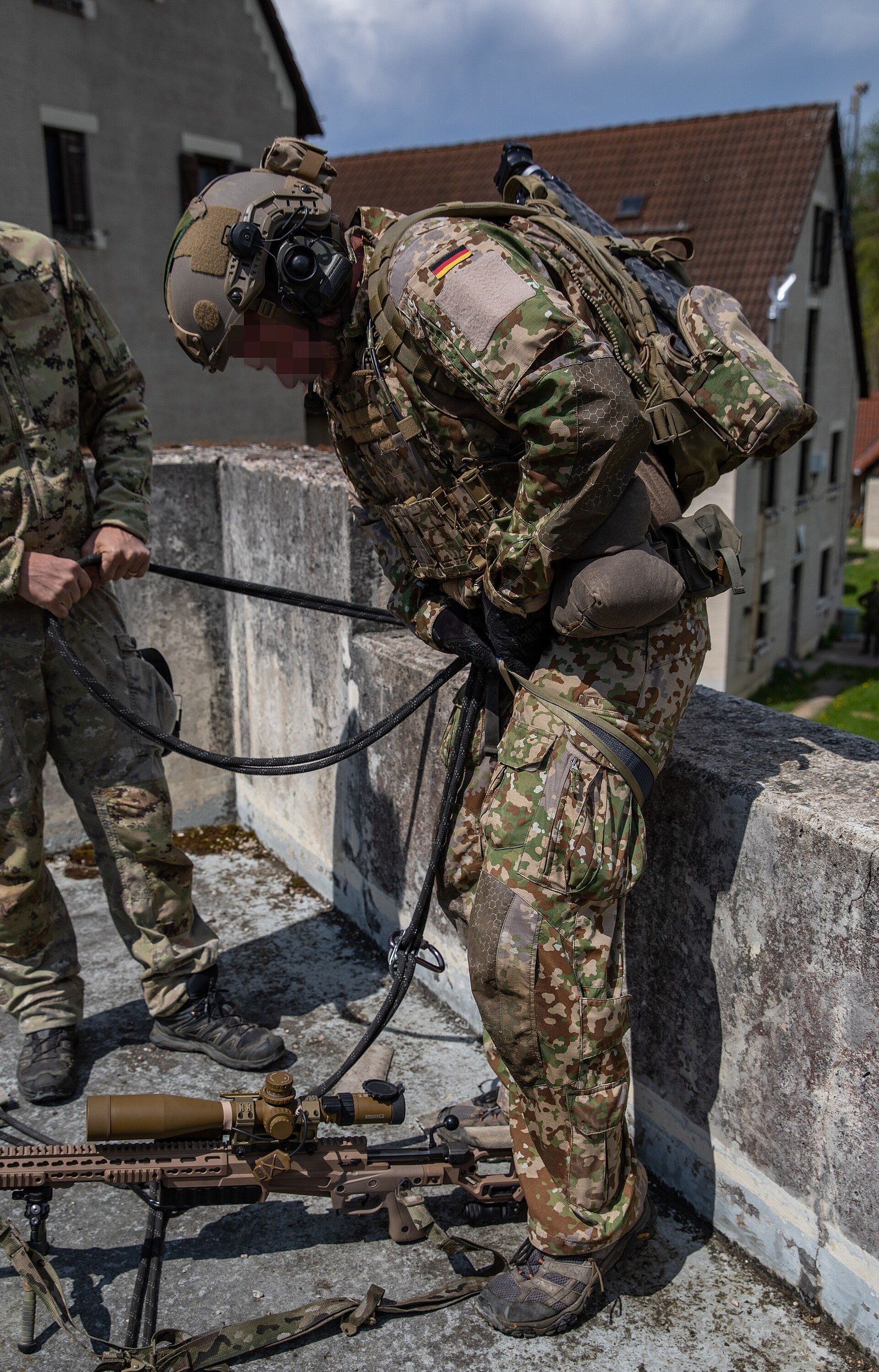 A German army sniper in Multitarn camouflage with a helmet, body armour and slung rifle preparing to rappel down the wall of a building, with a second soldier in a different camouflage at the left edge of the frame.