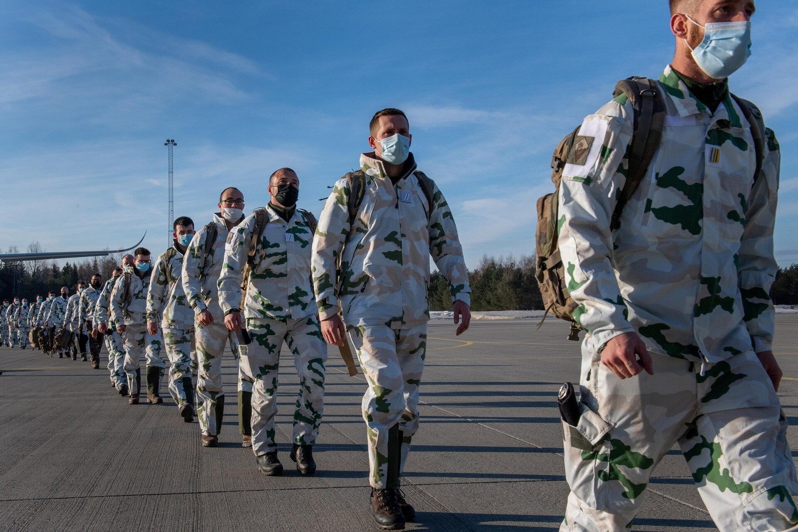 A column of French Alpine Hunters in the pale French alpine snow camouflage, with rucksacks and face masks, walking across an airfield apron.