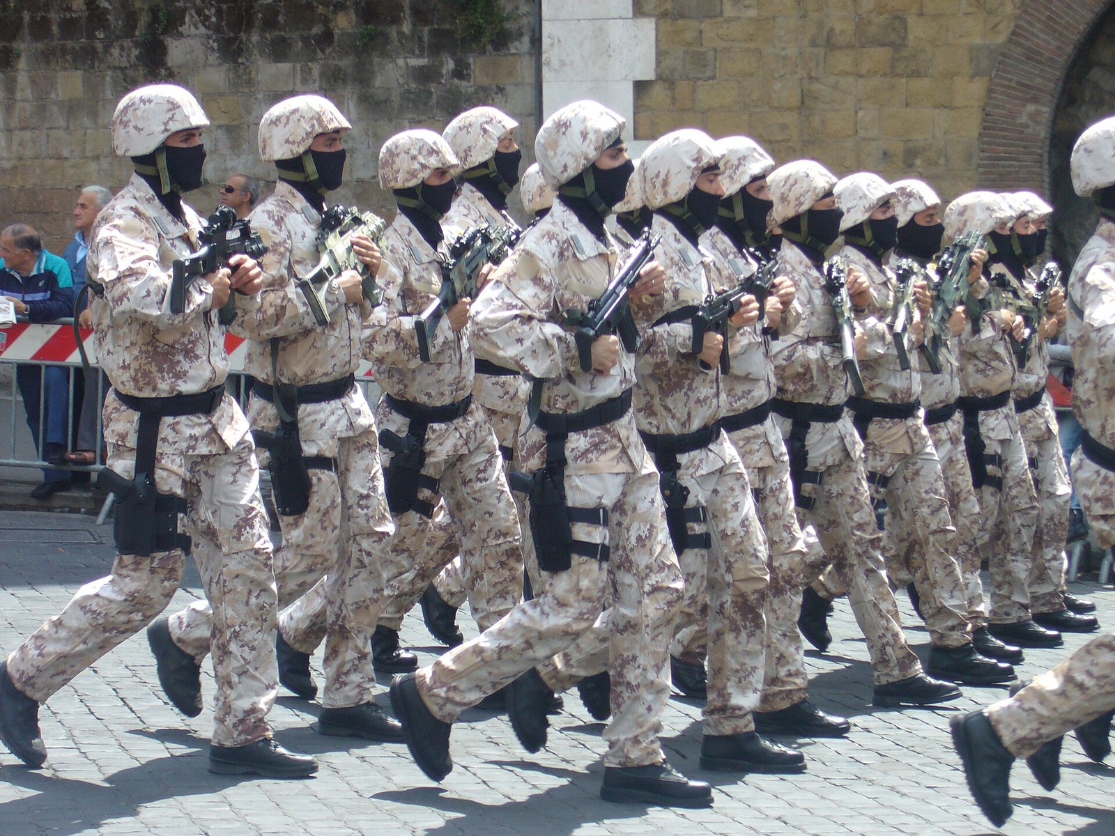 A column of Italian special-operations carabinieri marching in step through a cobbled city square in pale sand-and-reddish speckled camouflage, their helmet covers in the same print, their faces covered by black veils, holding black rifles.