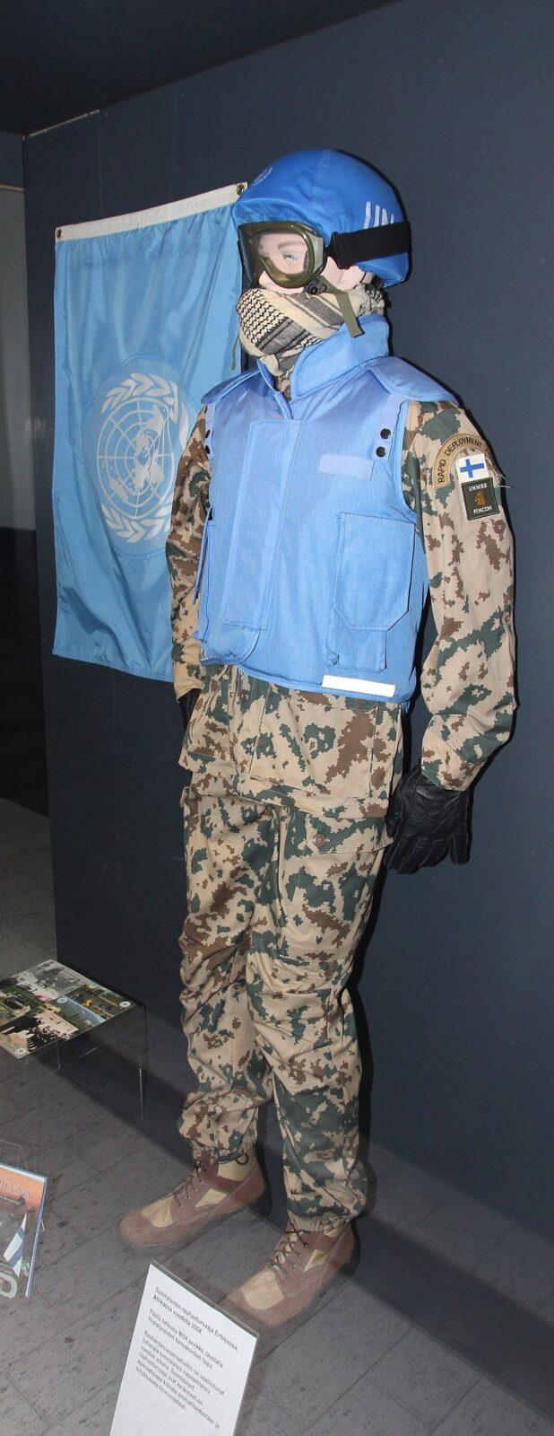 Museum display figure standing in a sandy desert camouflage uniform with green and brown blotches, covered by a blue United Nations vest and helmet, a UN flag on the wall behind, photographed through a glass case.