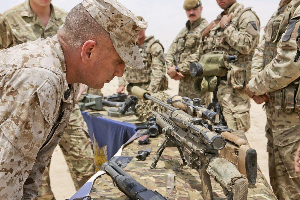 A senior U.S. Marine non-commissioned officer in desert digital camouflage examining a displayed rifle outdoors, with British service personnel in organic multi-tone camouflage standing around him.