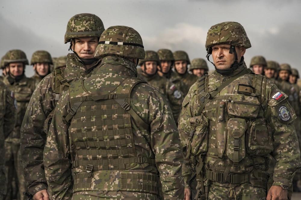 A group of Slovak soldiers in the blocky green camouflage of the vz 2007 Les print, in printed helmet covers and printed load-carrying vests, standing in loose formation at a training area.