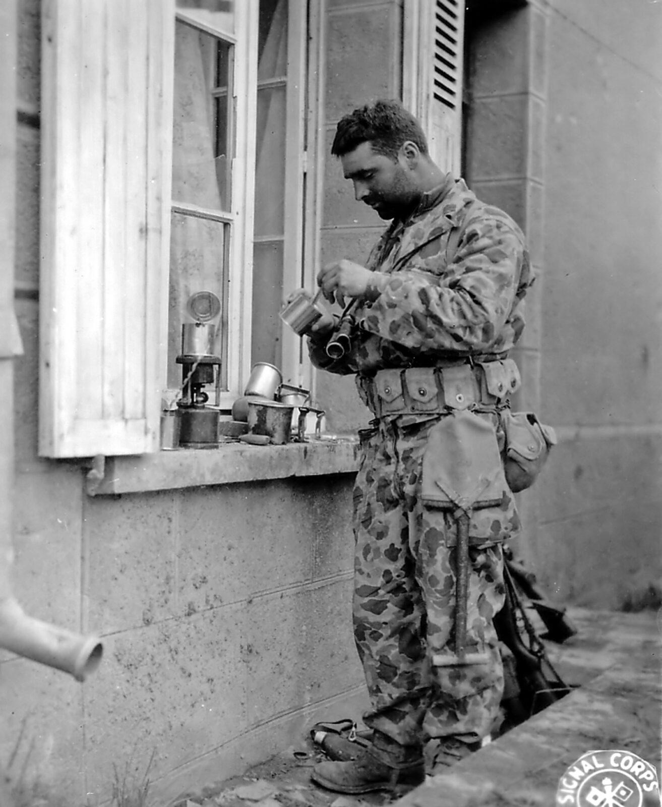A soldier in camouflage uniform standing at a windowsill, heating food on a small stove set on the sill beside a cup and mess tin, Normandy, July 1944.