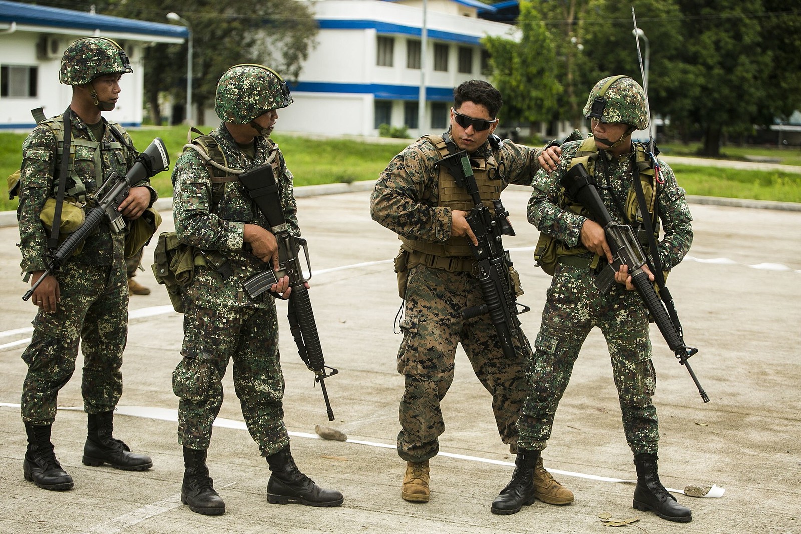 Three Marines in green square-block digital camouflage standing beside a fourth in a sand-coloured digital camouflage pattern during training on a paved apron.