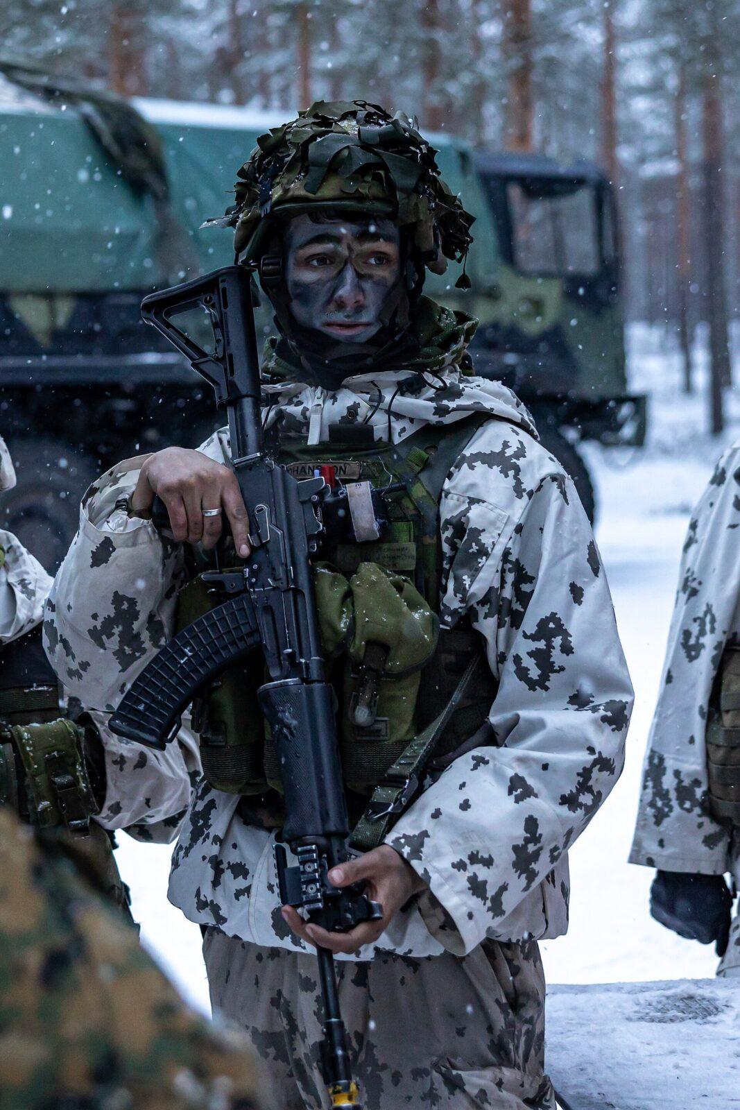 Soldier in falling snow holding a black assault rifle, wearing an off-white snow camouflage jacket with near-black blotches over greyish trousers, an olive load-bearing vest and a helmet covered with green foliage, a green truck and pine forest behind.