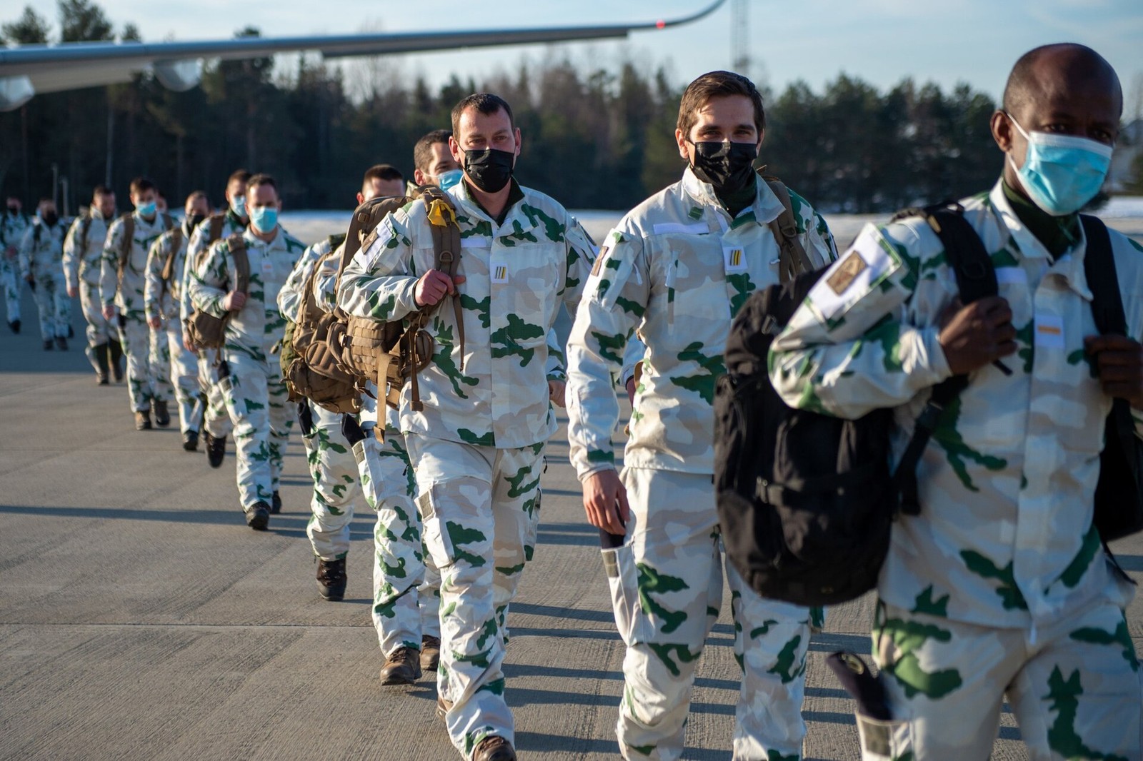 French Alpine Hunters in pale snow camouflage with green and salmon-pink shapes and face masks, carrying rucksacks past an aircraft on arrival in Norway.