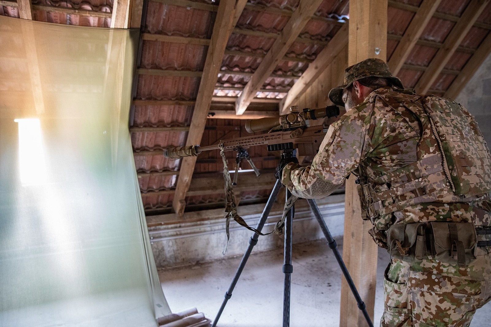A German special forces sniper in pale six-colour camouflage and a camouflage hat aiming a tripod-mounted scoped rifle from inside a loft space under a tiled roof.