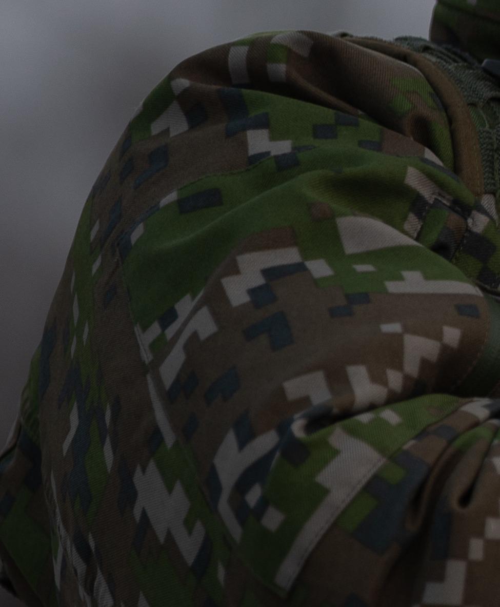 Detail of the shoulder and upper arm of a camouflage jacket showing quadrilaterals in several sizes, squares and long thin rectangles, in olive green, brown, khaki and dark green.