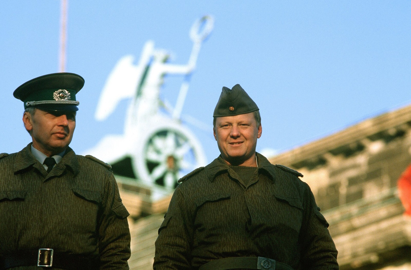 Two uniformed East German men, one in a peaked cap and one in a side cap, photographed against the Quadriga of the Brandenburg Gate on 14 November 1989, wearing fine vertical line camouflage field clothing