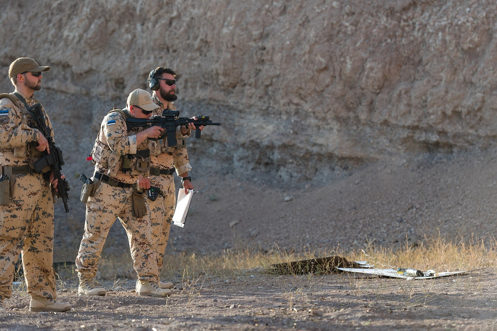 Estonian soldiers in the field wearing desert-pattern pixelated uniform and plain tan caps, seen from the front and side.