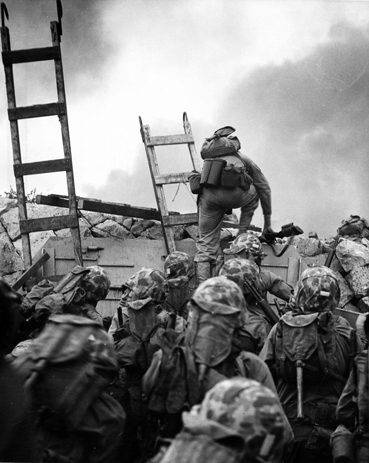 A marine officer climbing a wooden scaling ladder over a seawall during a beach landing, with other marines following behind.