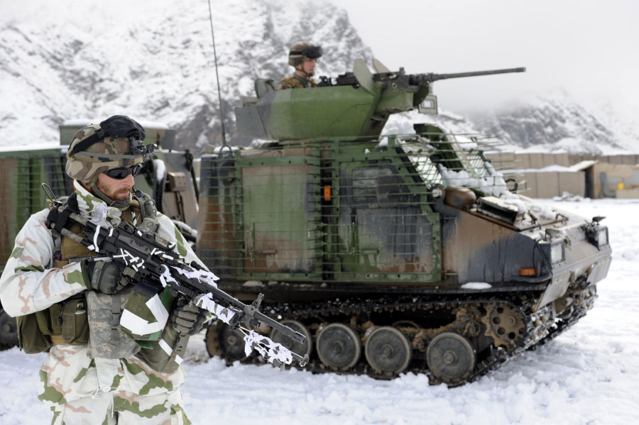 A soldier in pale Tundra camouflage — white ground with green and salmon shapes — with white strips wrapped over his weapon, standing in front of a green tracked armoured vehicle on snow, snow-covered rock faces behind.